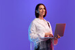 © master1305 - Beautiful woman, employee standing with laptop and smiling against purple studio background in neon light. Concept of business, employment, freelance, human emotions, fashion, lifestyle, ad