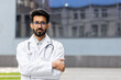 © Liubomir - Portrait of young confident and serious doctor, man thinking looking at camera with crossed arms, Indian man outside clinic in white medical coat.