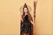 Hopeful girl dressed in witch costume holding broom isolated over beige background posing at halloween party keeps fingers crossed making wish desire.