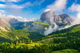 Fototapeta Psy - A vibrant depiction of the Sassolungo Massif and Gardena Valley in Dolomite Alps, Italy. The foreground showcases a lush green valley, while the background highlights a cloud-covered mountain peaks