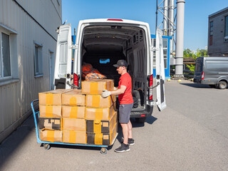 Wall Mural - Man unloads car. Loader guy near cart with cardboard boxes. Loading parcels into truck. Man loads goods into minivan. Warehouse trolley with boxes. Loader near building with storage units