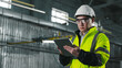 © ihorvsn - Asian engineer looks at screen of tablet. Worker wearing yellow vest, glasses and hard hat. Male stands on background of workflow. Man at workplace.