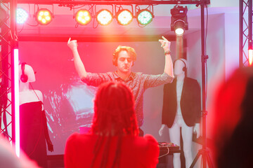 Young man dj in headphones standing on stage with spotlights at nightclub concert. Musician with closed eyes raising hands while enjoying electronic music at disco party in club