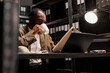 Leinwandbild Motiv African american cop drinking coffee and reading csi report at night time. Detective sitting at workplace desk, holding tea mug and studying crime case file in dark office room