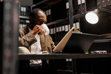 African american cop drinking coffee and reading csi report at night time. Detective sitting at workplace desk, holding tea mug and studying crime case file in dark office room