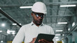 © ihorvsn - Serious thoughtful African-American hard-working male employee manufacturer factory manager engineer using tablet. Man wearing glasses white safety hardhat nice shirt.