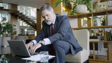 Canvas Print - Businessman sitting at table in hotel lobby, restaurant or cafe, working on laptop computer. Mature age man in business casual, confident happy smiling