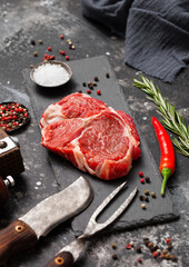 Wall Mural - Raw Rib eye fillet on stone board with fork and knife,salt and pepper with rosemary on black kitchen table background.