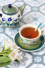 A beautiful green cup and saucer with tea on a table with a tea pot and white flowers. Selective focus.