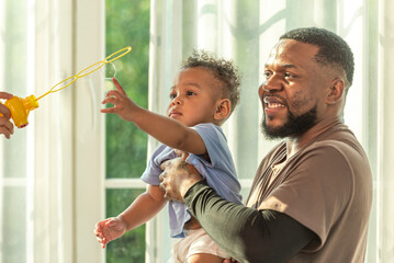 Wall Mural - Happy african black parents dad father throw baby son on piggyback and neck riding teasing fun near window. Black baby son and daddy enjoy teasing kiss cheek and neck riding at window light