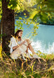 Beautiful young woman sitting under the tree hugging dog near the lake enjoying the silence and beauty of nature. Mindfulness, pet love and unwind yourself concept