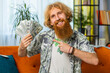 Successful happy rich Caucasian redhead man counting money cheering up with high profits, lottery game win, satisfied of wealth income earnings salary, on home room couch full of dollar cash banknotes