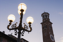 A Clock Tower And Illuminated Lamp Post In Piazza Delle Erbe At Dusk; Verona, Italy