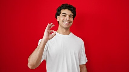 Poster - Young latin man doing ok gesture smiling over isolated red background