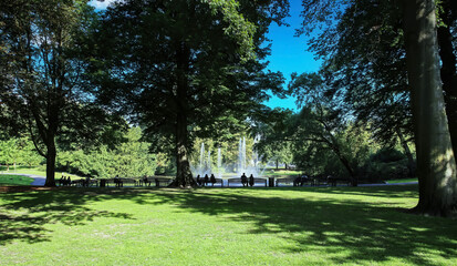 Breda, Netherlands - August 30. 2023:  Unrecognizable people sitting on benches in shade of trees at beautiful pond,  water fountain in green city park Valkenberg on sunny summer day