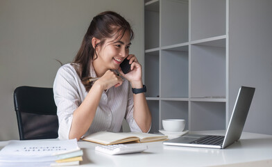 Asian businesswoman talking on mobile phone working on laptop in modern office. Happy business woman talking on mobile phone while analyzing weekly schedule in laptop