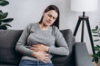 Upset sad woman holding belly and feeling abdominal menstrual pain or bowel and digestion problems. Unhealthy young caucasian female suffering from stomach ache sits on couch. Food poisoning concept