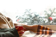 red mug in hands against the background of a window decorated with fir branches. cozy dreaming on winter holidays
