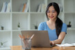 © crizzystudio - Asian woman sitting happily at work smiling relaxing and chatting with friends on social media search for information on laptop take notes in a notebook while sitting on a desk in his office at home
