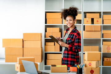 Wall Mural - Young African woman prepares parcel boxes and checks online orders for delivery to customers on laptop. Shipping and online shopping concepts