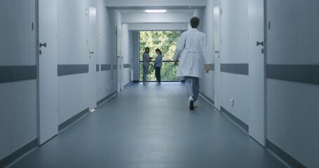 Wall Mural - Clinic corridor: Doctors and professional medics walk. Nurse with digital tablet comes to elderly woman standing near window. Medical staff and patients in modern hospital or medical center hallway.