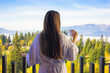 Brunette girl long hair from back in bathrobe standing on balcony. Elegant woman is drinking tea or coffee from a white cup enjoying morning.