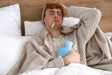 Poster - Ill young man with hot water bottle measuring temperature in bedroom