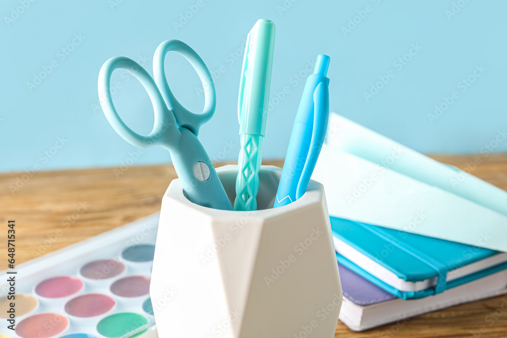 Holder with pens, scissors, paints and notebooks on wooden table, closeup