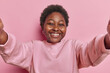 © WHstudio Leushin N - Attractive dark skinned chubby young woman smiles broadly keeps arms outstreched poses for making selfie tries to find striking pose for photo being in good mood isolated over pink background