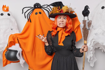 People and October holiday concept. Funny woman makes grimace wears wizard hat and dress poses with broom near orange ghost surrounded by creepy creatures prepares for Halloween celebration.