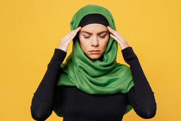 Young arabian asian muslim woman wear green hijab abaya black clothes put hands on head rub temples have headache solated on plain yellow background. People uae middle eastern islam religious concept.