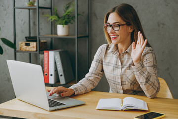 Young smiling happy fun successful employee business woman wearing shirt casual clothes glasses sit work at office desk with pc laptop computer waving hand talk speak. Achievement career job concept.