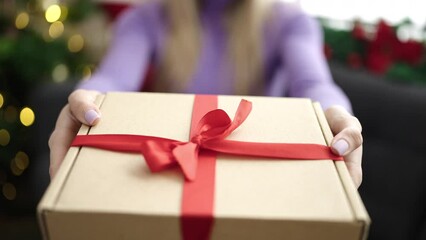 Poster - Young blonde woman holding gift sitting on sofa by christmas tree at home