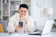 © Liubomir - Asian mature man sitting alone at home on sofa in living room cinema, man received online notification message with bad news on phone, upset browsing internet pages using app on smartphone.