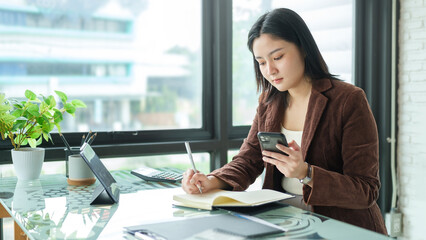 Wall Mural - Beautiful asian businesswoman sitting at her workplace and using smart phone.