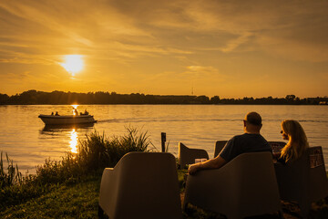 A mature couple enjoy sunset drinks by the waterside watching nature as the day comes to an end. Evening drinks at lakeside by fresh water in natural beauty on summer evening with boat passing by.