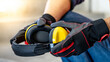 © Summer Paradive - Male worker hand with black and red protective gloves holding yellow safety ear muffs or ear protectors preparing to wear on his head. Equipment for high noise reduction