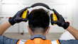 © Summer Paradive - Back view of male worker with reflective orange vest and protective hand gloves pulling yellow safety ear muffs or ear protectors when wearing on his head. Equipment for high noise reduction