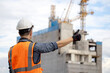 © Summer Paradive - Construction worker man with orange reflective vest and safety helmet showing thumbs up at unfinished building structure and tower crane. Male site engineer showing hand gesture sign for approval