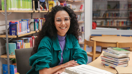 Portrait of a charming smiling Oriental girl with curly hair wearing glasses sitting at a school desk in a classroom or school library, backdrop of bookshelves smiling directly looking at the camera.