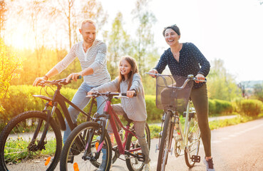 Wall Mural - Smiling father and mother with daughter during summer outdoor bicycle riding. They enjoy togetherness in the summer city park. Happy parenthood and childhood or active sport life concept image.