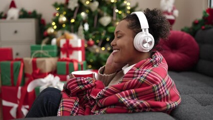 Poster - African american woman listening to music drinking coffee sitting by christmas tree at home