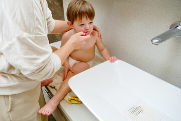 Baby and his mother are brushing their teeth in the bathroom. Kid aged two years (two-year-old boy) Mom helps child brush her teeth with a toothbrush