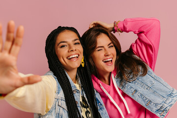 Wall Mural - Two joyful young women in colorful wear embracing and smiling against pink background together