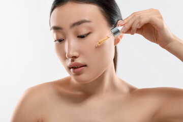 Wall Mural - Beautiful young woman applying cosmetic serum onto her face on white background, closeup