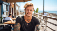Beautiful Attractive Young Man With Blonde Hair And Body Tattoos. Enjoying Outdoors At A Lively Boardwalk Lined With Shops And Restaurants, Emphasizing A Carefree Spirit
