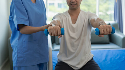 Handsome man exercising with dumbbells and stretching straps smiling in hospital department. Attractive nurse is helping him, orthopedic doctor helps do physical therapy, physical therapy in the clini