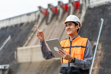 Wall Mural - Professional asian maintenance engineer man with safety helmet in construction site dam with hydroelectric power plant and irrigation. Manager engineer man working with tablet at project big building.