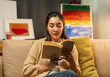 © Chanakon - Asian beautiful woman sitting reading book on cozey couch sofa in living room with warm light at night. People female stay home part time relax at home.