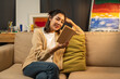 © Chanakon - Asian beautiful woman sitting reading book on cozey couch sofa in living room with warm light at night. People female stay home part time relax at home.